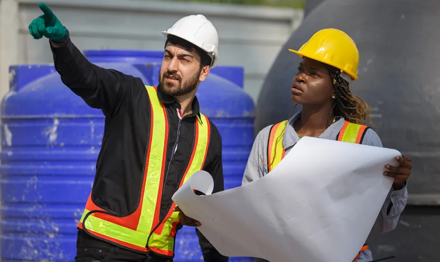 Two construction workers wearing safety vests and hard hats discuss plans at a worksite. One holds blueprints while the other points ahead, with large blue and gray tanks in the background.