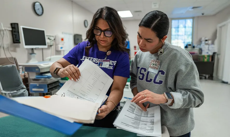 Two women in a medical setting look over documents together; one wears glasses and a purple shirt, the other has dark hair pulled back and wears a gray LSCC sweatshirt. Medical equipment is visible in the background.