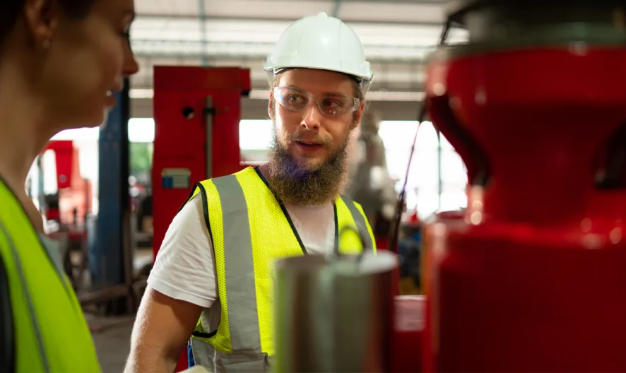 Two workers wearing high-visibility vests and hard hats stand in a factory, having a discussion near red industrial machinery. One worker, with a beard and glasses, looks at the other person attentively.