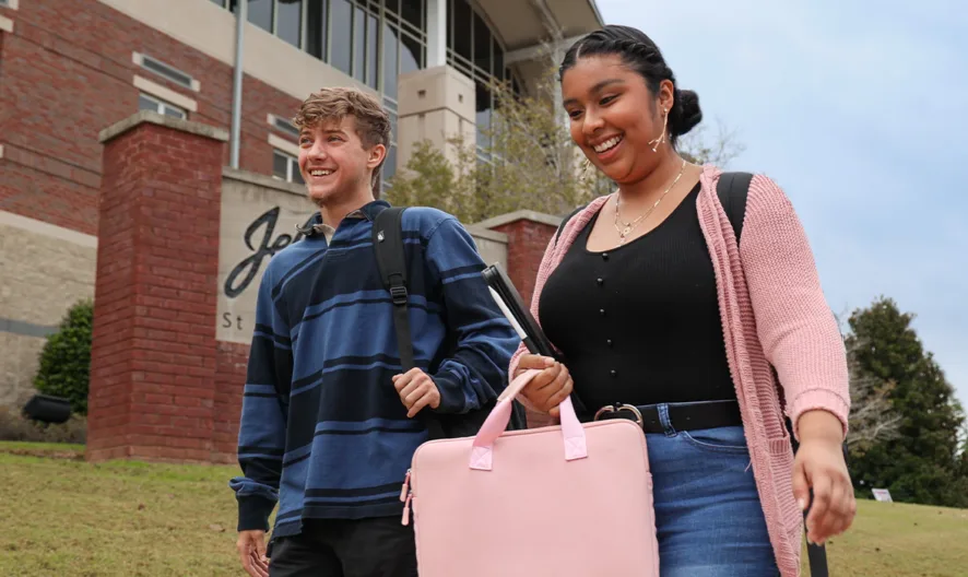 Two smiling college students, one carrying a pink bag, walk outside a campus building on a cloudy day. The buildings brick and stone exterior is visible in the background, along with greenery and a campus sign.