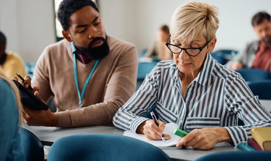 A woman with short gray hair and glasses writes in a notebook at a classroom desk while a man next to her looks on. Other adults are seated in the background, suggesting a learning environment.