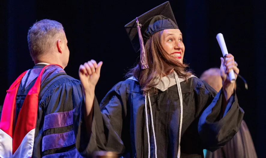A smiling graduate in a cap and gown holds up a diploma and celebrates on stage, while another person in academic regalia stands nearby with their back turned.