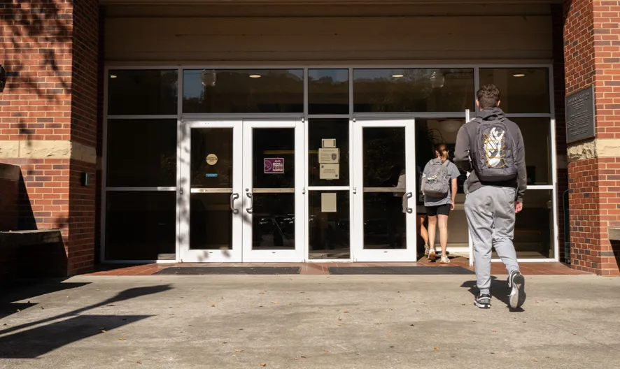 Two students with backpacks approach the entrance of a brick building with glass double doors on a sunny day.