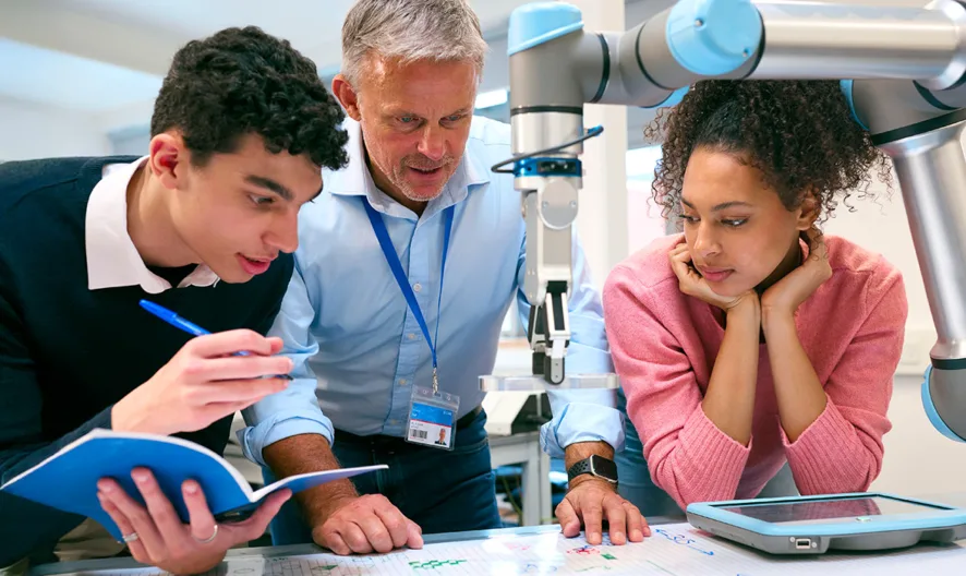 A teacher and two students work together at a table with a robotic arm. The students take notes and observe while the teacher explains, creating a collaborative learning environment with technology.