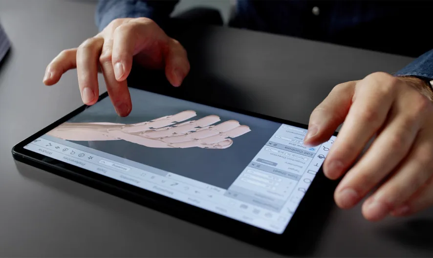 A person uses a tablet with a stylus to design a digital 3D model of a robotic hand, with graphs and papers on the desk nearby.
