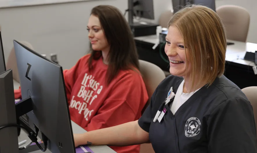Two women sit at computer desks in a classroom. One wears a red shirt and types, while the other, in dark scrubs with a badge, smiles while working. Both are focused on their monitors.