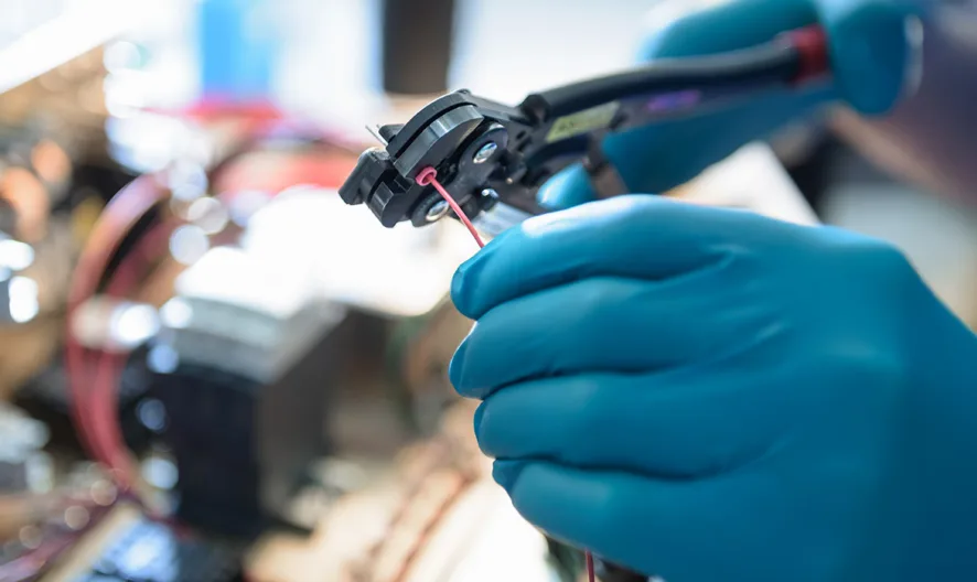 A person wearing blue gloves uses a wire stripper to remove insulation from an electrical wire, with electronic components and cables visible in the background.