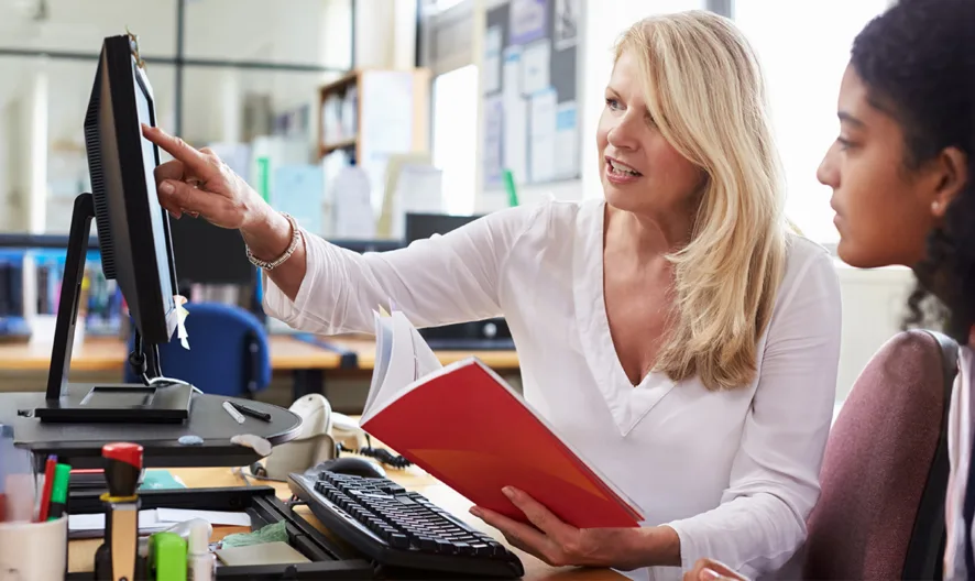Two women sit at a desk in an office. One is pointing at a computer screen while holding a red notebook, and the other is attentively watching. Office supplies and shelves are visible in the background.