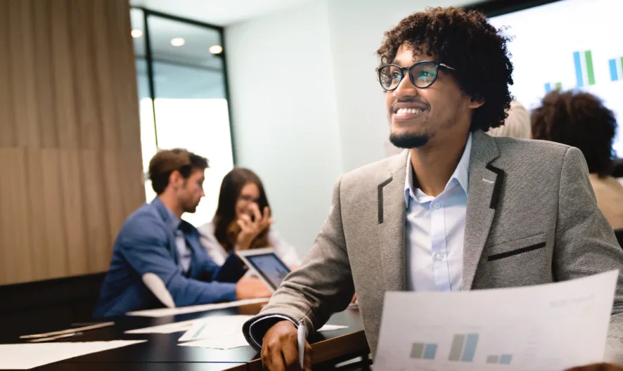A smiling man in a blazer holds a document with graphs in a modern office. Three colleagues work together in the background, some using laptops, with a large screen displaying charts on the wall.