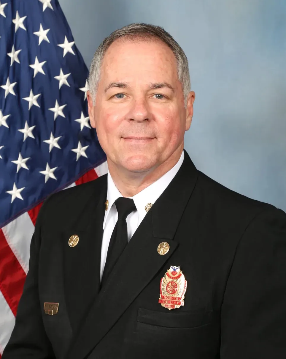 A middle-aged man in a formal black uniform with fire department insignia stands in front of an American flag, smiling slightly.