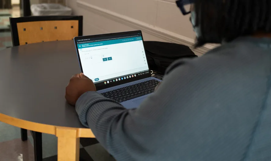 A person wearing a mask works on a laptop at a round table in a hallway with checkered tile flooring. The screen displays a website with text and graphics.