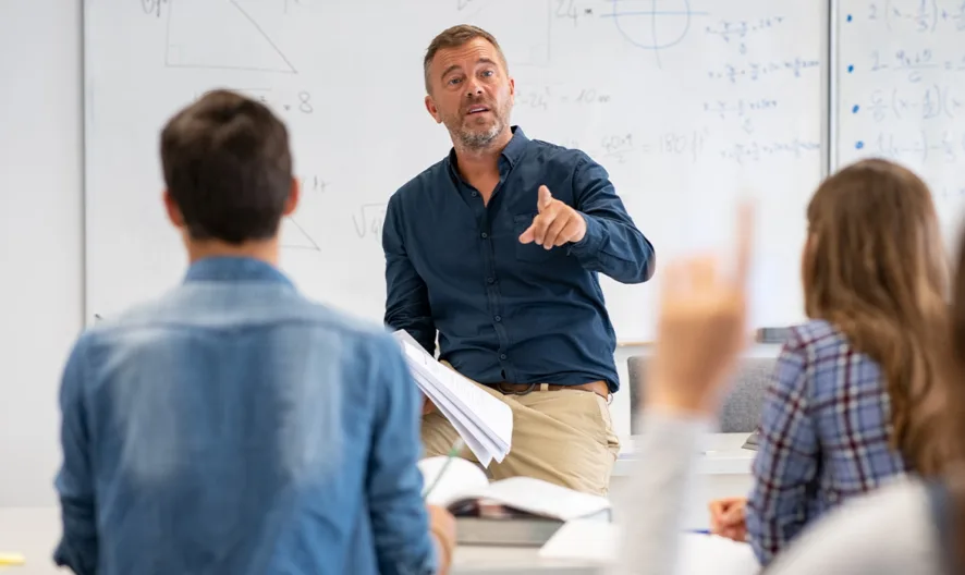 A teacher sits on a desk at the front of a classroom, pointing while speaking to students. Several students face the teacher, one with a hand raised. Mathematical equations are written on the whiteboard behind them.