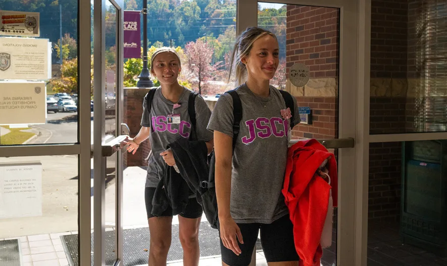 Two young women wearing matching JSU t-shirts and shorts walk through glass doors into a building, carrying bags and papers, with sunlight and a parking lot visible outside.