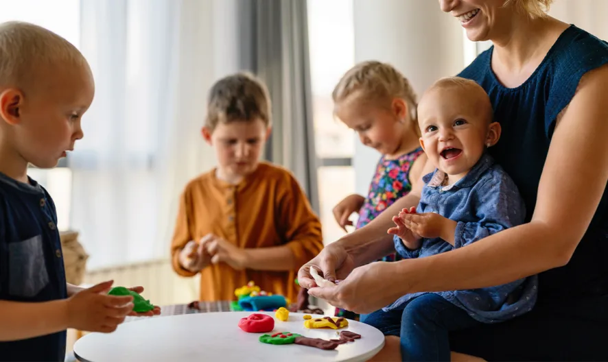 A woman sits with a smiling baby on her lap at a small table, while three young children play with colorful modeling clay in a bright, cozy living room.