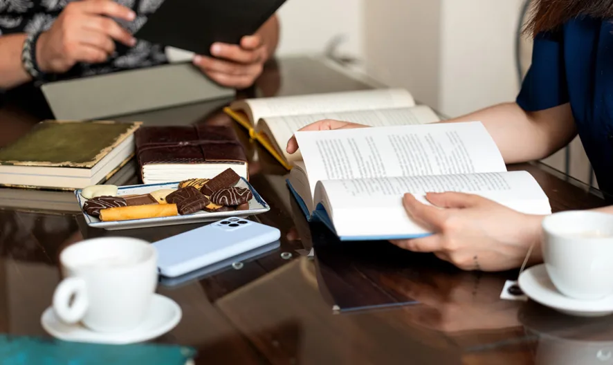 Two people sit at a wooden table with books, a tablet, coffee cups, a plate of cookies and chocolates, and a smartphone, engaged in reading and conversation.