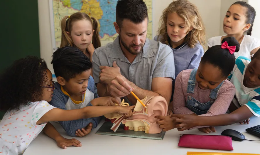 A teacher sits at a table with six young students, showing them a model of a human ear. The children eagerly point and look at the model, appearing engaged and curious. A map and computer are visible in the background.