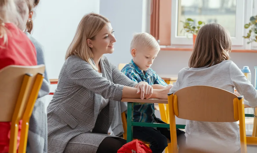 A teacher sits beside young children at classroom tables, assisting them with activities. Sunlight comes through the windows, and potted plants decorate the sill. The scene is warm and focused on learning.