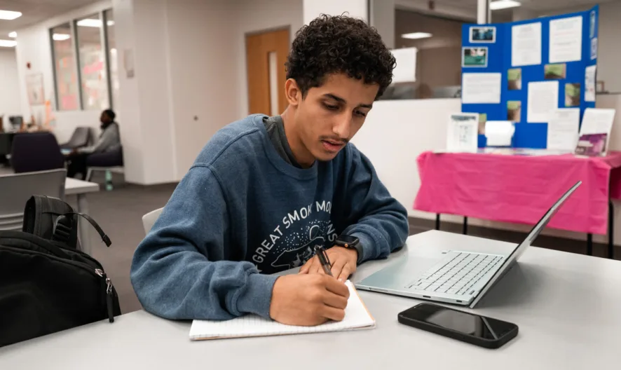 A young man sits at a table in a library, writing in a notebook with a laptop open beside him. A phone and backpack are also on the table. Bulletin boards and other students are visible in the background.