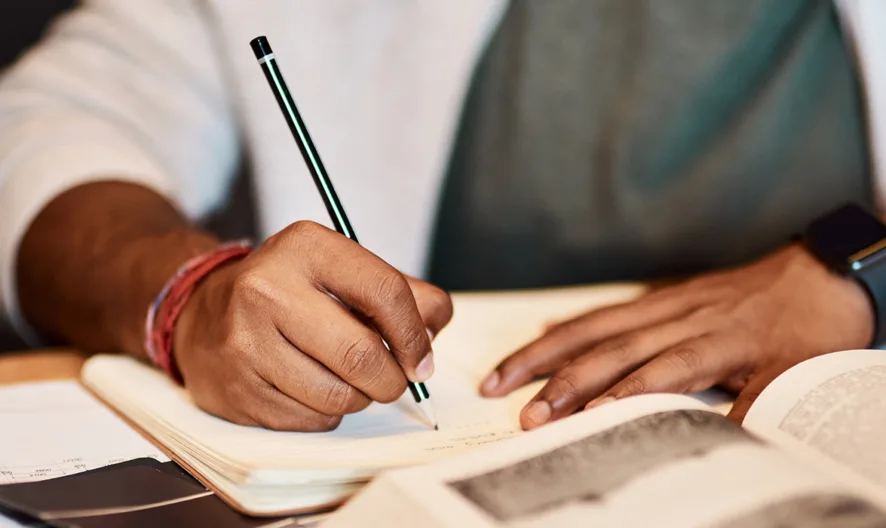 A person wearing a smartwatch writes in a notebook with an open textbook and documents on the desk, suggesting studying or working.