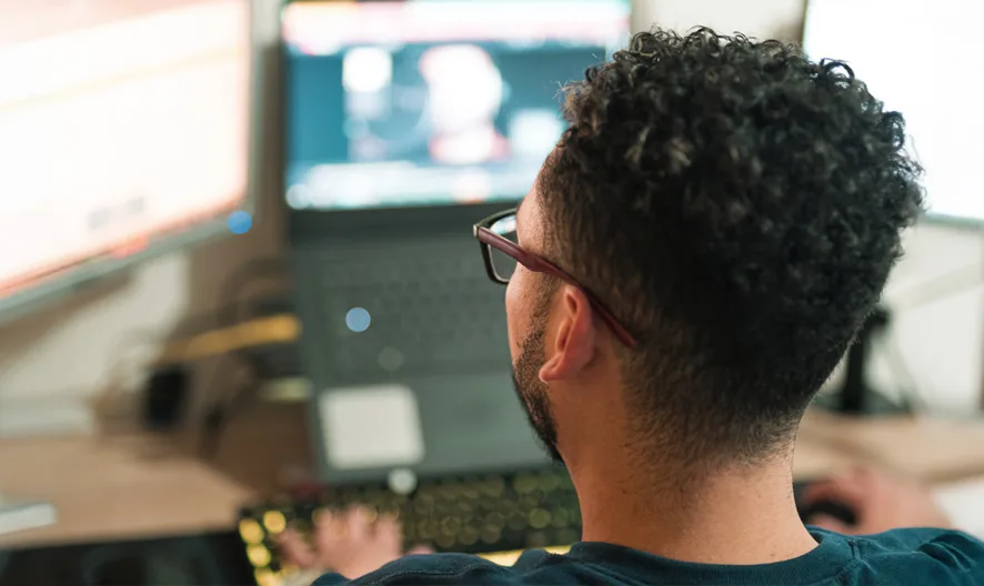 A person with short curly hair and glasses is sitting at a desk, working on a laptop and two large computer monitors in a home office setting. The screens display various digital content.