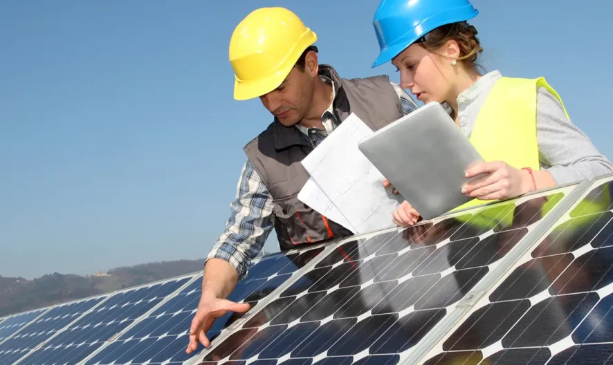 Two workers wearing safety helmets and vests inspect solar panels outdoors; one holds documents and a tablet while the other points at the panels, with mountains and blue sky in the background.