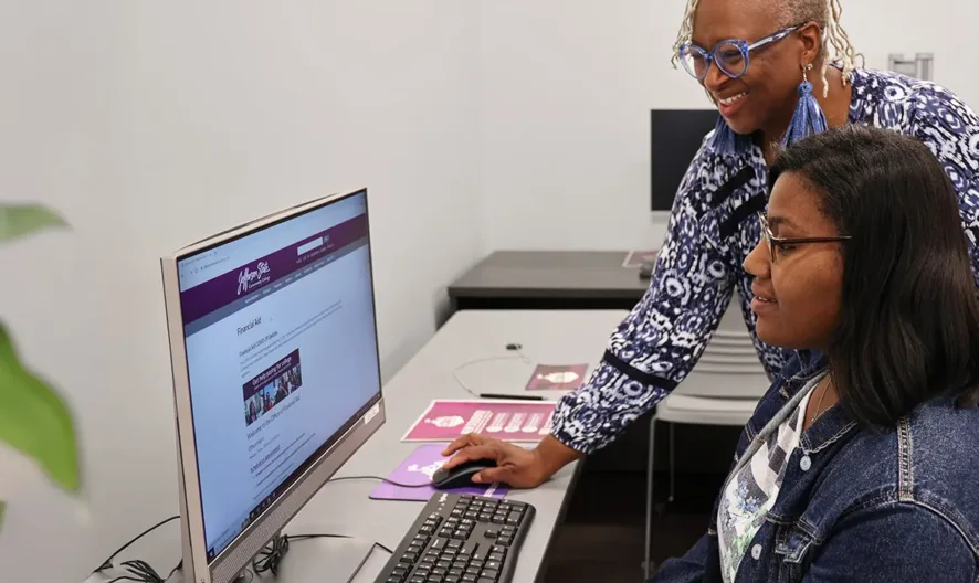 Two women are using a desktop computer together in a classroom. One woman, standing, is smiling and assisting the seated woman. Another person sits at a computer in the background. A plant is visible in the foreground.