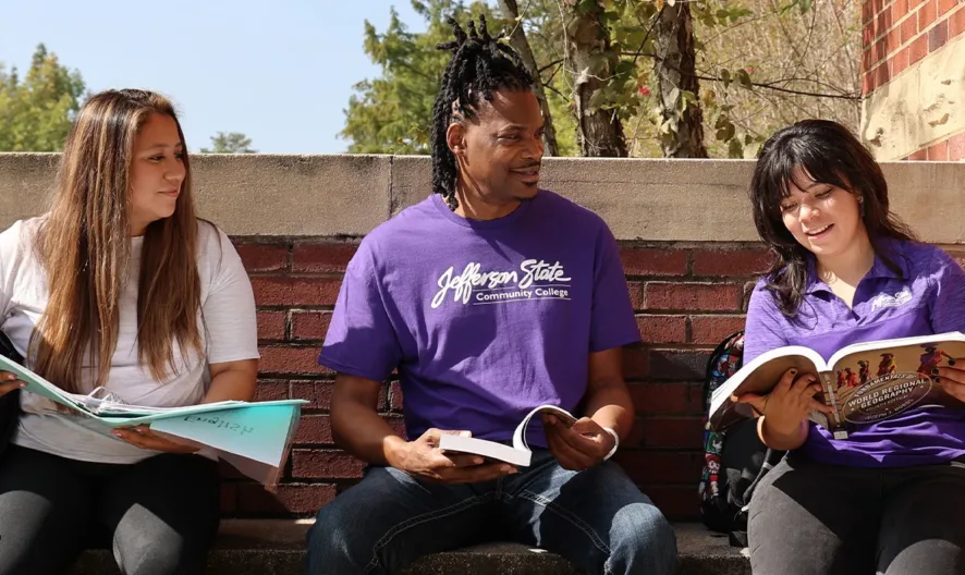 Three students sit on an outdoor brick bench, smiling and reading books together. Two women and one man, all casually dressed, appear to be studying and enjoying a sunny day on campus.