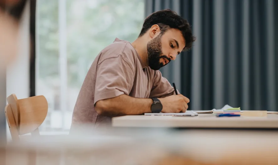 A man with a beard sits at a desk, concentrating as he writes in a notebook. There are papers and stationery on the desk, and the background features large windows and blue curtains.