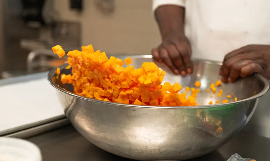 A person in a white chef’s coat mixes a large bowl of diced orange vegetables, likely squash, in a commercial kitchen setting.