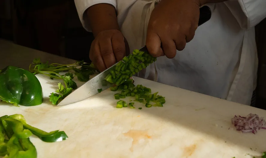 A person in a white chefs coat chops green bell pepper on a white cutting board, surrounded by sliced vegetables including onion and more peppers.