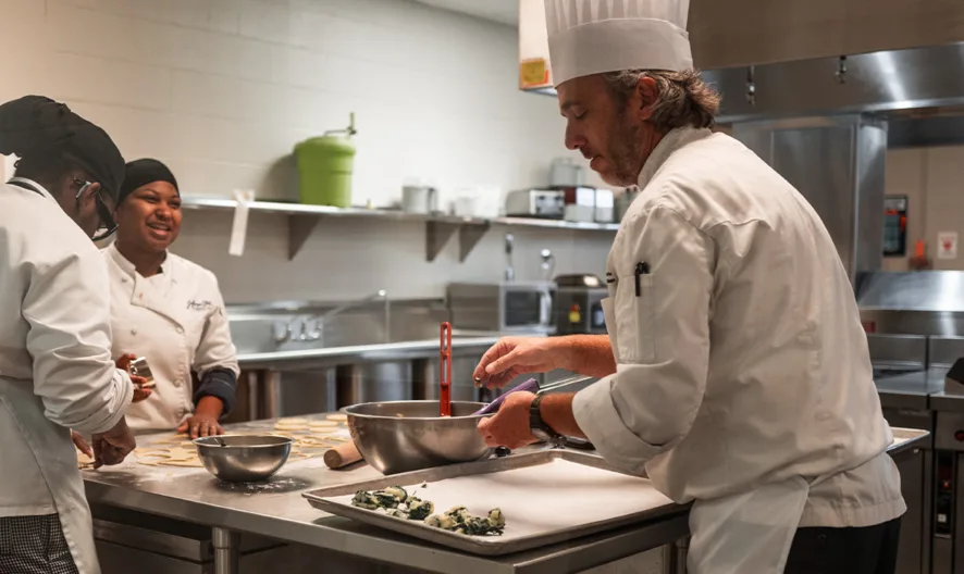 Three chefs in white uniforms and hats work together in a commercial kitchen. One chef mixes ingredients in a bowl, while the others prepare food nearby on the counter, with ovens and utensils in the background.
