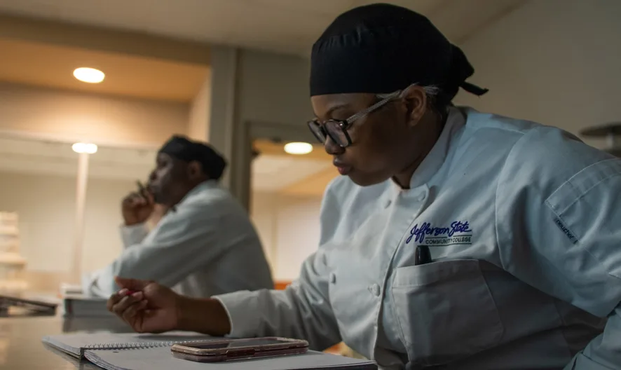 Two chefs in white uniforms and black head coverings sit at a table; one writes in a notebook while the other looks ahead thoughtfully. A smartphone and more notebooks are on the table. Shelves and cakes are visible in the background.