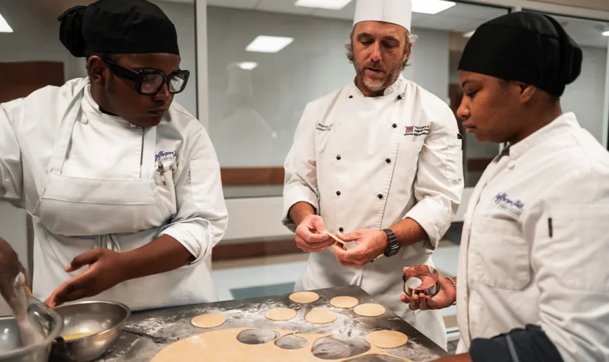 Three chefs in a kitchen prepare dough; one chef mixes ingredients, another shapes dough circles, and a chef in a white hat supervises and offers guidance.