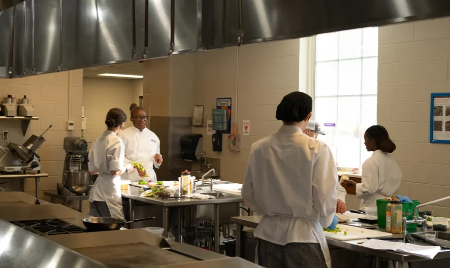 Several people in white chef uniforms work at stainless steel counters in a bright commercial kitchen, preparing food and using various kitchen tools under large ventilation hoods.
