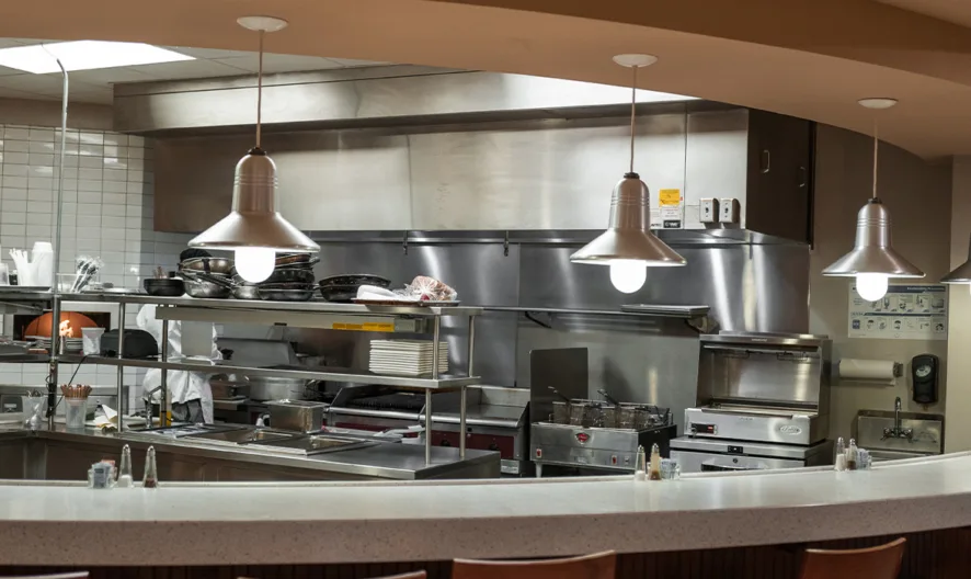 A clean, modern commercial kitchen with stainless steel appliances, hanging pendant lights, a curved counter with stools, and shelves holding kitchen supplies and equipment.