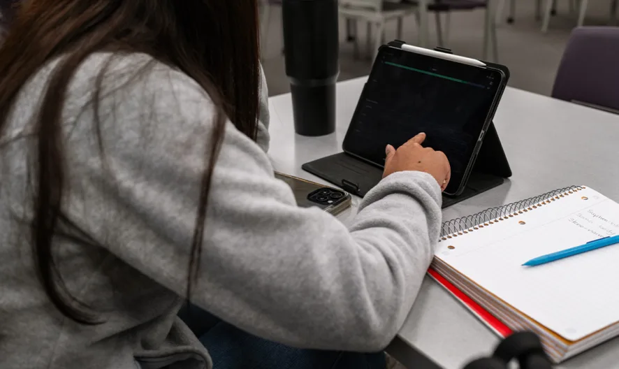 A person with long hair, wearing a gray sweater, sits at a table using a tablet. An open notebook with a blue pen rests on the table in front of them, and the setting appears to be a classroom or study area.
