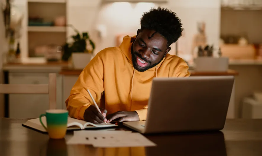 A man in a yellow hoodie sits at a table, smiling as he writes in a notebook beside his open laptop. A colorful mug and some papers are also on the table, and the background shows a warmly lit kitchen.