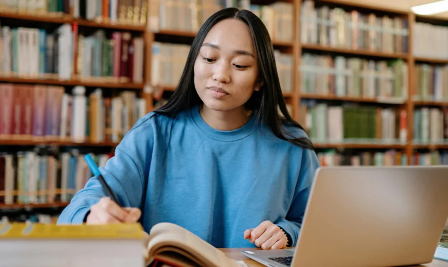 A young woman in a blue sweater studies at a library table, taking notes in a notebook with an open textbook and a laptop in front of her, surrounded by bookshelves filled with books.