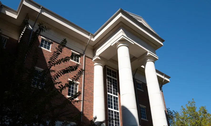 A low-angle view of a red brick building with tall white columns and large windows, set against a clear blue sky with some tree branches visible in the foreground.