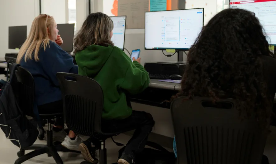 Three people sit at desks using desktop computers in a classroom or lab setting. They are focused on their screens, and one person is also looking at a smartphone. The room has several empty chairs and computer stations.