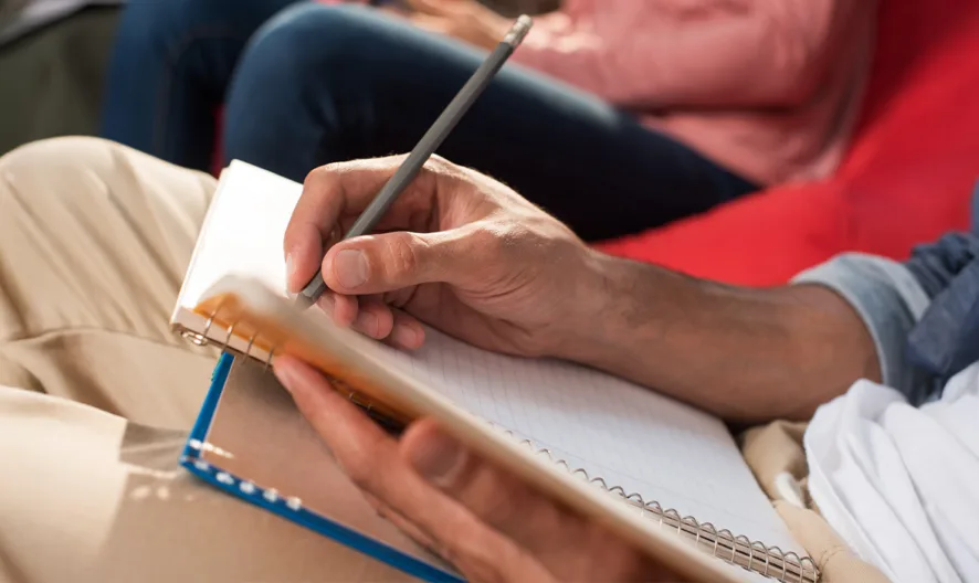 Close-up of a person sitting and writing in a spiral notebook with a pencil. Another person is seated in the background, slightly out of focus. The setting appears casual and relaxed.
