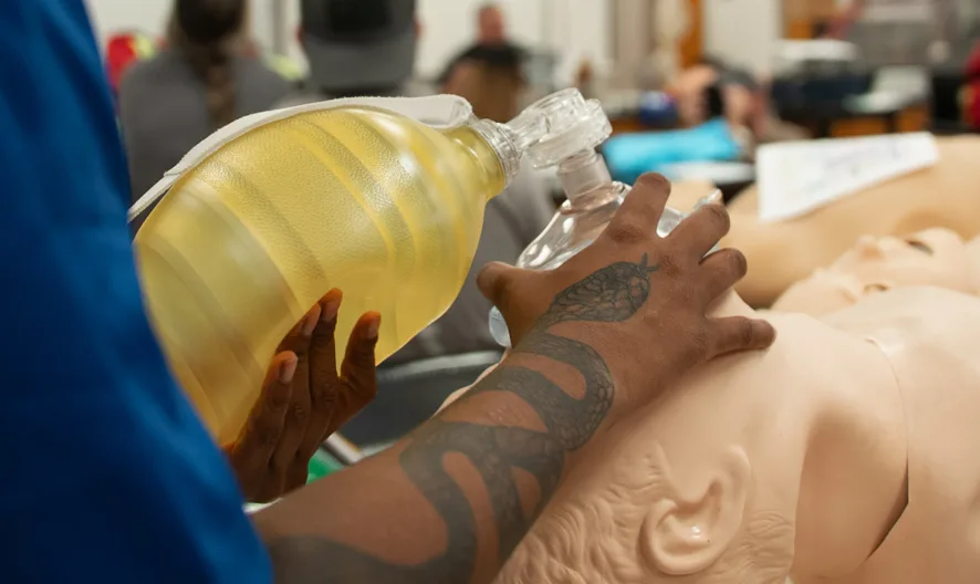 A person with a tattooed arm uses a manual resuscitator to practice CPR on a medical training mannequin in a classroom setting.