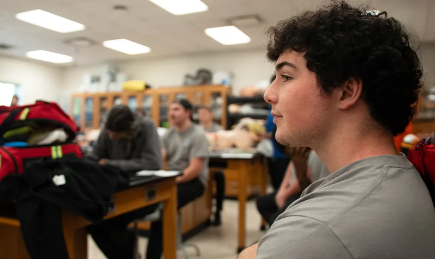 A student with curly hair sits in a science classroom, surrounded by classmates at lab tables. The focus is on the student in the foreground, while others and academic equipment are visible in the background.