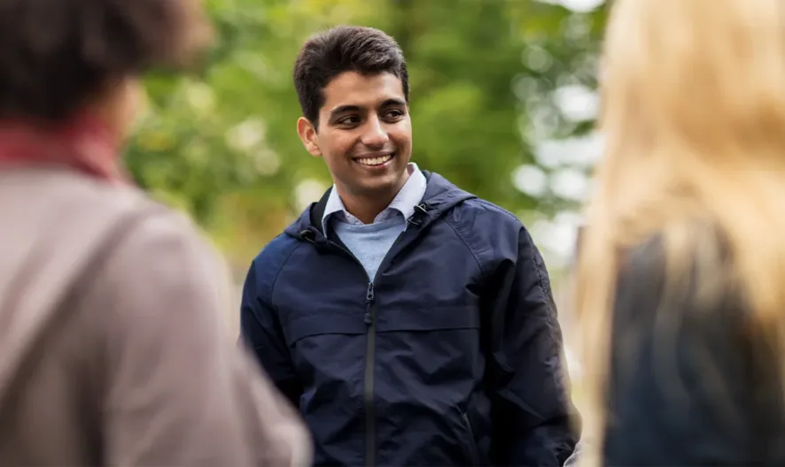 A young man wearing a navy jacket smiles while talking to two people outdoors. The background shows green trees, suggesting a park or natural setting. The two people seen from behind have curly and straight hair.