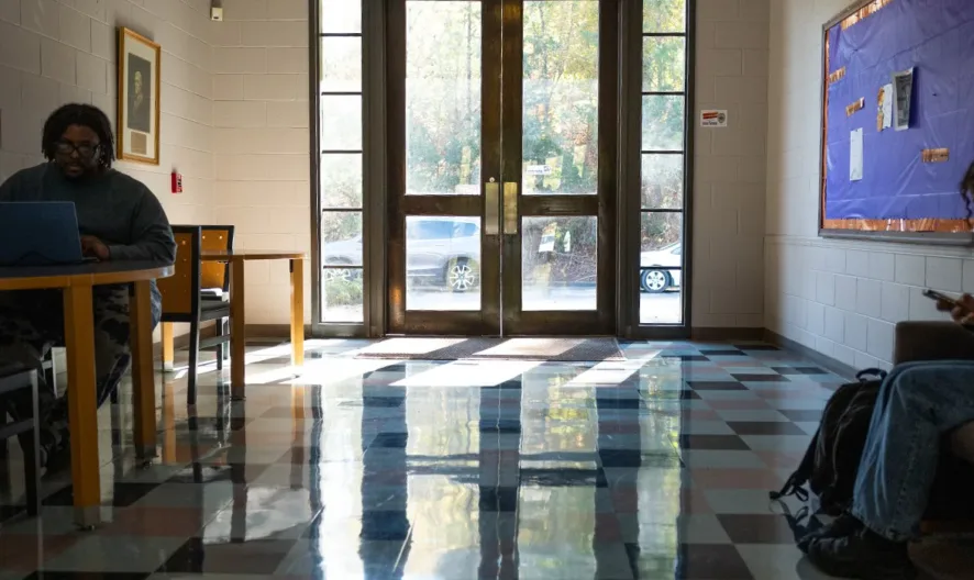 Two people are in a sunlit hallway; one sits at a table using a laptop, the other sits on a chair using a phone. Light streams in through glass doors, casting reflections on the checkered floor.