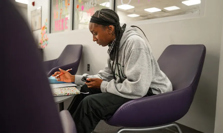 A person sits in a purple chair, focused on writing in a notebook while holding a phone. They wear a gray hoodie and black pants, with classroom or office surroundings visible in the background.