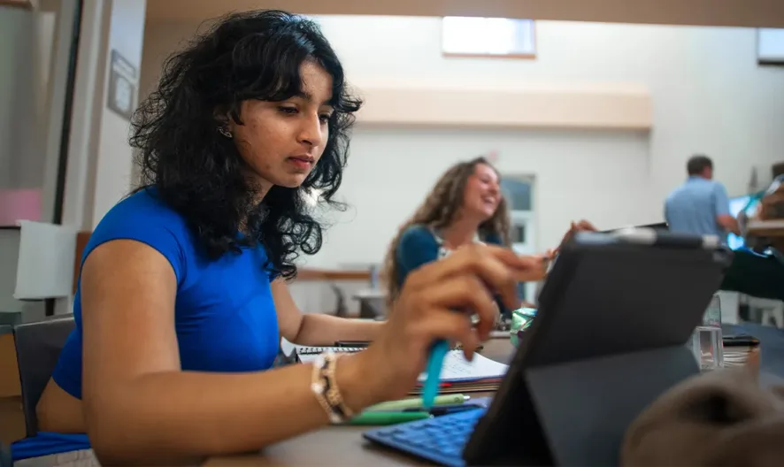 A woman in a blue shirt sits at a desk using a tablet and stylus, focused on her work. In the background, two people are having a conversation and another person is standing near shelves.