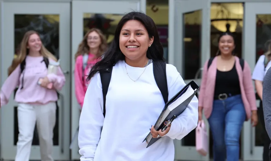 A diverse group of high school students smiling and walking out of a school building, carrying backpacks and books, with glass doors and classmates in the background.