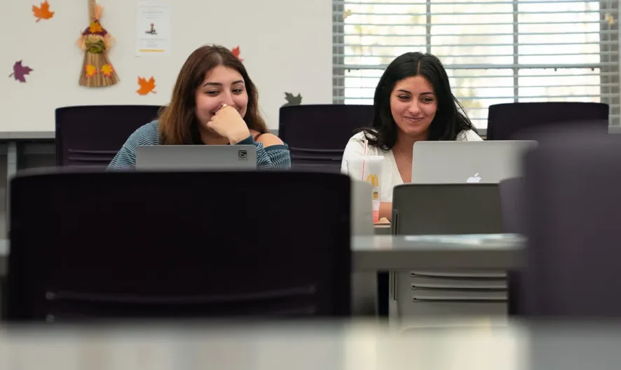 Two women sit side by side at a table in a classroom, smiling and working on laptops. The room is sparsely decorated with fall-themed wall art and mostly empty chairs and tables.