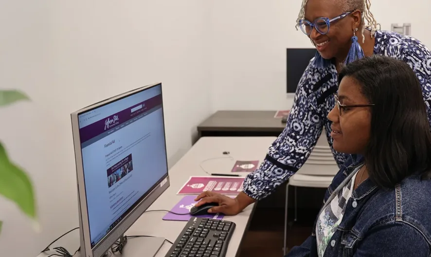 Two women work together at a computer in a classroom. One is seated using the mouse, while the other stands beside her, smiling and assisting. Another person works at a computer in the background.